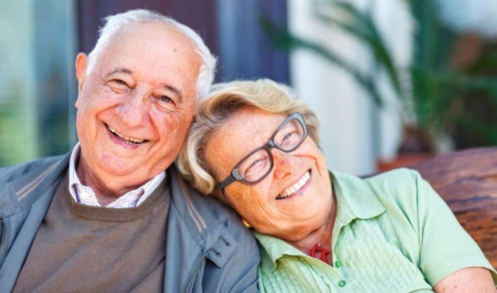 An elderly couple sits closely together, both smiling warmly at their fortuna. The man is wearing a gray jacket and brown sweater, while the woman sports glasses and a green shirt. They seem to be enjoying their time outdoors at an elder care home with a blurred background.