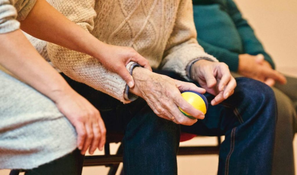 In an elder care home, two people sit closely together. One, in a beige sweater, holds a colorful stress ball while anothers hand rests gently on their arm, offering support. Nearby, Fortuna stands partially visible in a green top.