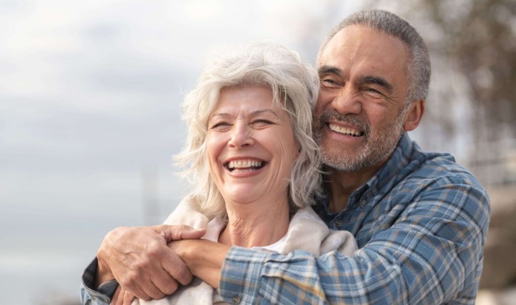 An older couple stands outside, with a man embracing a woman from behind. Both are smiling broadly, exuding joy and contentment. The background hints at a natural setting near their elder care home, Fortuna, where happiness and care intertwine seamlessly.