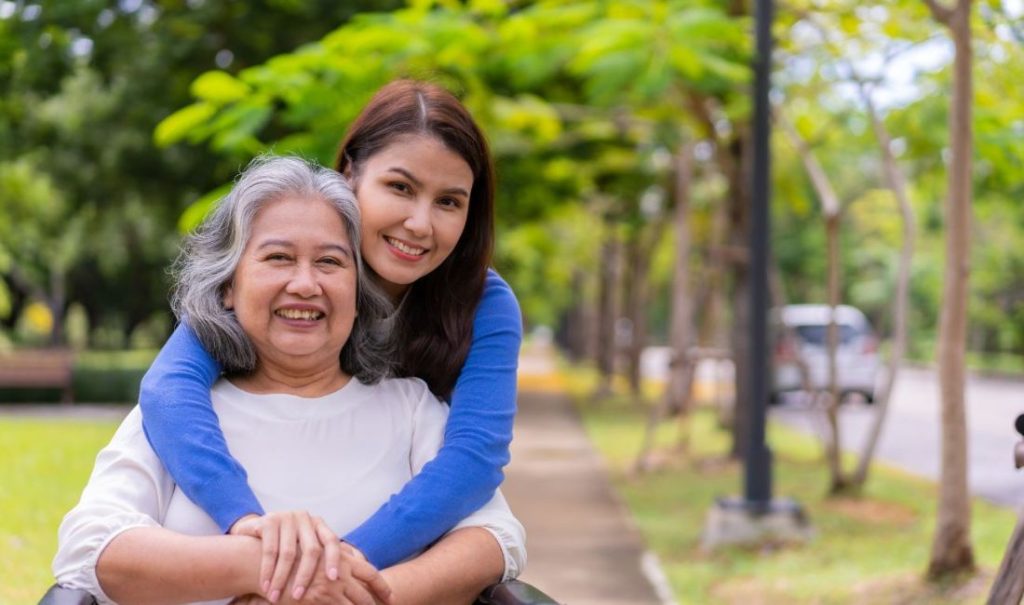 A smiling young woman in a blue shirt hugs an older woman from behind. They are outdoors at Fortuna Park, where the green trees and winding path create a serene backdrop, reminiscent of walks they enjoyed during visits to the elder care home.