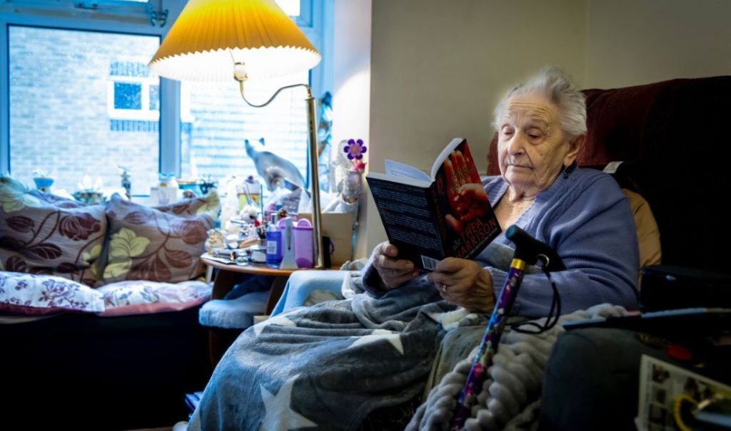 An elderly person wearing a robe is sitting in a cozy, dimly lit living room, reading a book. A walking cane rests beside them. The room is furnished with cushions, a lamp, and small tables holding various items, including a potted plant.