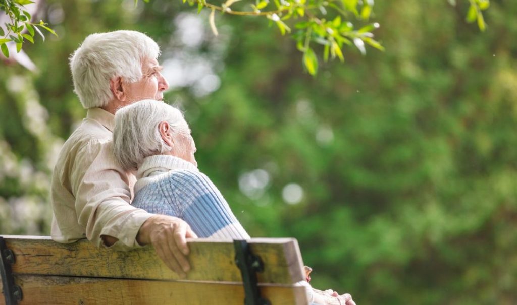 An elderly couple sits closely on a wooden bench at Fortuna Elder Care Home, enjoying a peaceful moment together. The man has his arm around the woman, who rests her head on his shoulder. Lush green foliage surrounds them, creating a serene outdoor setting.