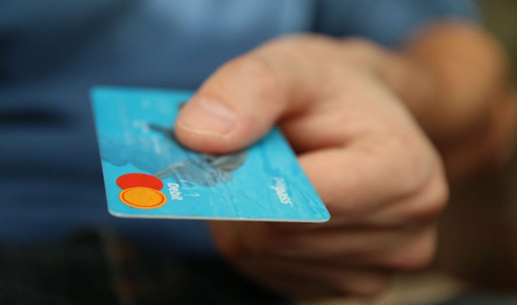 A person at the elder care home holds out a blue debit card with red and yellow circle logos and Debit printed on it. The blurred background adds a sense of fortuna to the moment, highlighting ease and accessibility in daily transactions.
