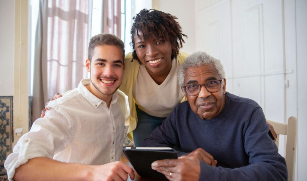 Three people, two younger and one older, are gathered around a table with a tablet at Fortuna elder care home. The younger ones are smiling, while the older person looks at the camera with a gentle expression. Natural light filters through a window behind them.