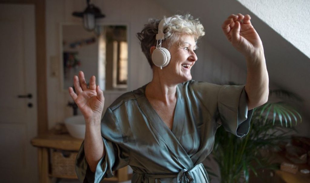 An older woman with short gray hair dances joyfully at the elder care home Fortuna, wearing headphones. Dressed in a green robe, she is in a cozy room with a wooden table and a plant in the background, smiling and raising her arms.