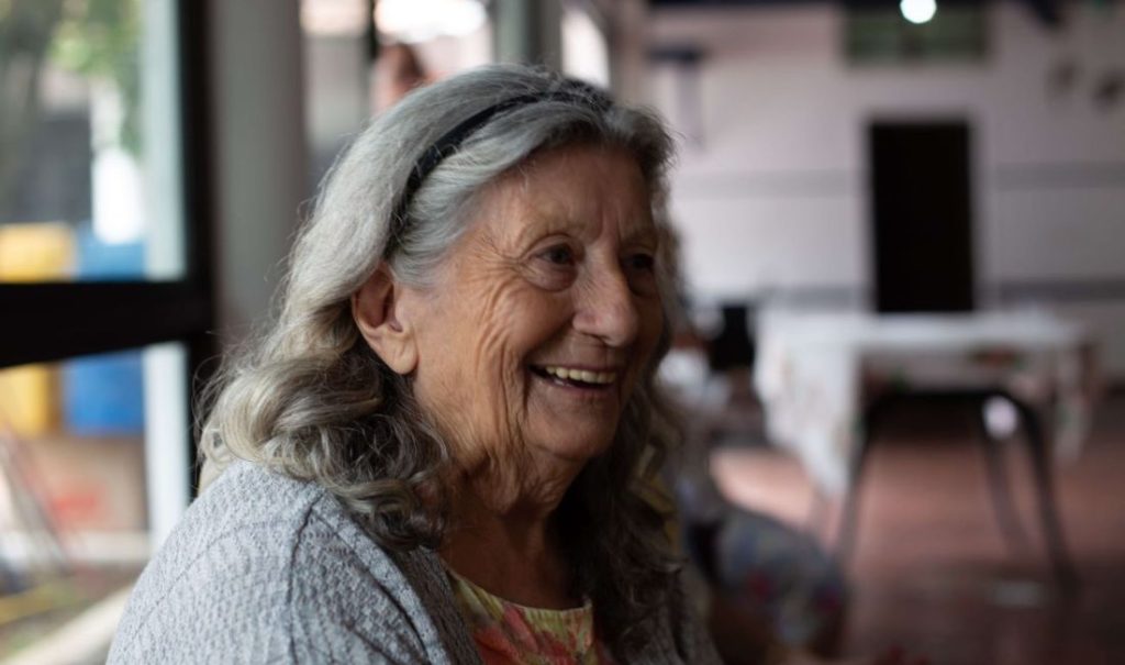 An elderly woman with long gray hair and a warm smile, wearing a headband and patterned blouse, sits comfortably indoors at Fortuna Elder Care Home. Blurred chairs and tables fill the background, as natural light gently streams in from a side window.