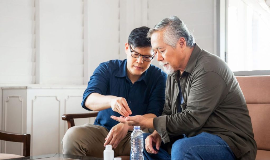 Two men sit on a sofa at a table in an elder care home. The younger man, wearing glasses and a blue shirt, hands pills to the older man in green. A closed water bottle and pill bottle rest on the table, embodying Fortunas watchful embrace of their daily routine.
