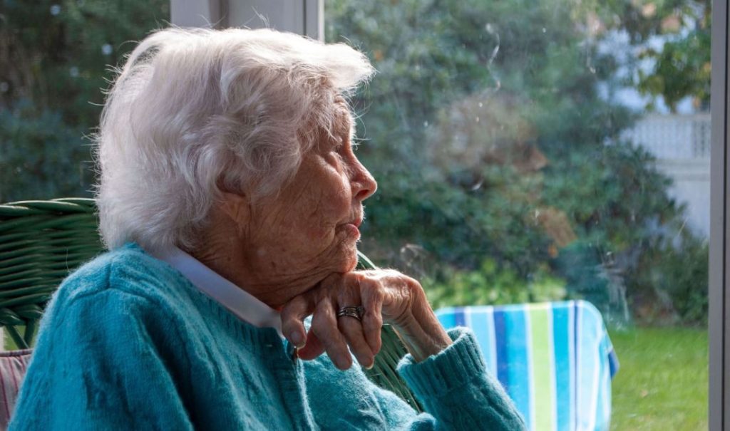 An elderly woman with white hair, clad in a turquoise sweater, sits in a wicker chair by the window of Fortuna Care Home, gazing thoughtfully outside. The background is adorned with lush greenery and a striped towel.